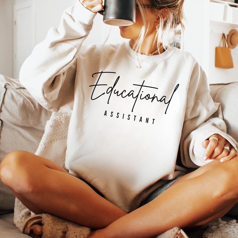 May include: Cream-colored sweatshirt with the words "Educational Assistant" in black script. The sweatshirt is a crew neck style and is made of a soft material. The person in the image is holding a black mug.