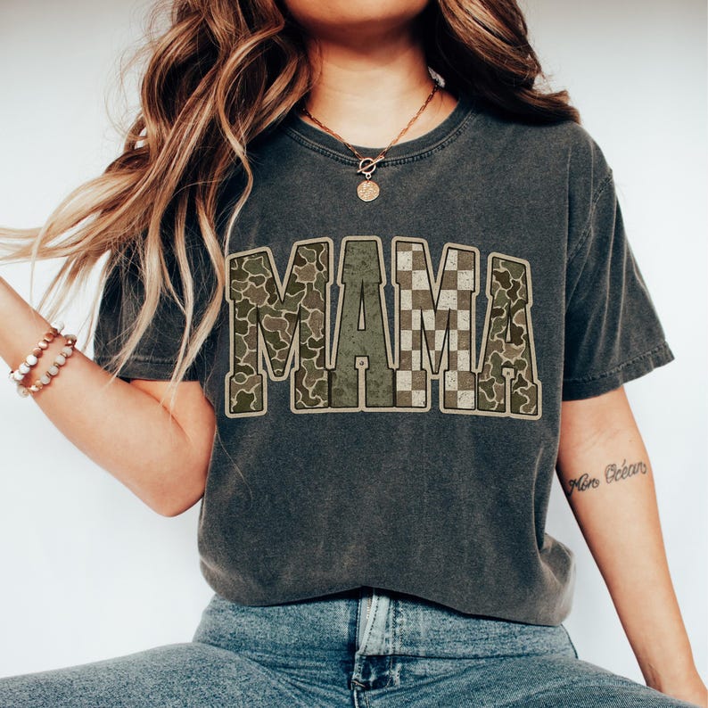 Puede incluir: Camiseta gris oscuro con la palabra "MAMA" en un dise&ntilde;o de camuflaje y cuadros. La camiseta tiene un aspecto vintage y desgastado, con un corte relajado. La palabra "MAMA" es el punto focal, con las letras rellenas con una mezcla de patrones de camuflaje verdes, marrones y beige.
