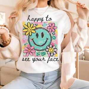May include: White t-shirt with the text "happy to see your face". The shirt features a turquoise smiley face surrounded by colourful flowers in yellow and pink. The t-shirt is part of a casual clothing collection.