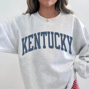 May include: A pale grey sweatshirt featuring the word "KENTUCKY" in navy blue block lettering. The top has a crew neck and long sleeves.