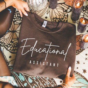 May include: A brown sweatshirt with the words "Educational Assistant" in white script. The sweatshirt is folded and held by hands, with a patterned background featuring butterflies and candles.