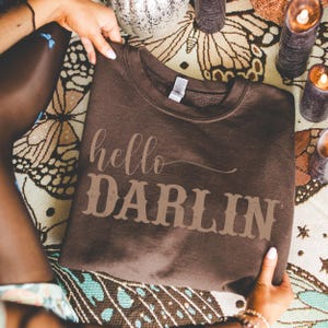 May include: A brown sweatshirt with the words "hello DARLIN" printed in a lighter brown color. The sweatshirt is held by a person, and the background includes candles and a decorative cloth.