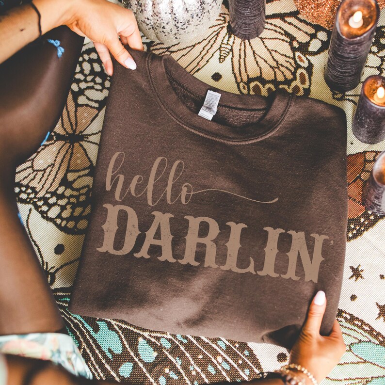 May include: A brown sweatshirt with the words "hello DARLIN" printed in a lighter brown color. The sweatshirt is held by a person, and the background includes candles and a decorative cloth.
