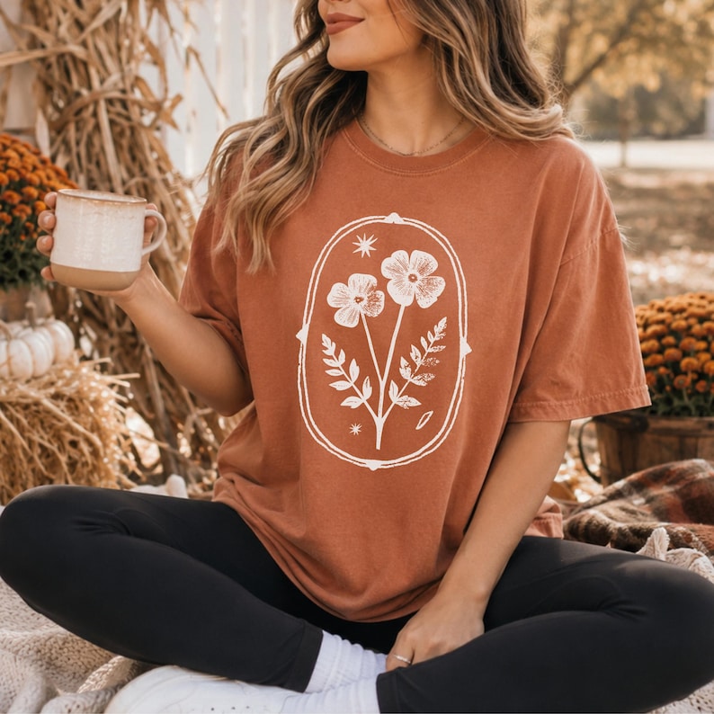 May include: A rust-colored short-sleeve t-shirt featuring a white graphic print of two flowers and leaves enclosed within an oval frame with small star accents. The shirt has a crew neckline and a relaxed fit, paired with solid black leggings.