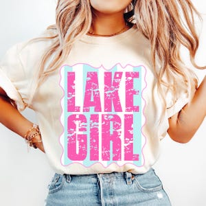 Puede incluir: Camiseta color crema con el texto "LAKE GIRL" en letras rosas desgastadas. El texto est&aacute; sobre un fondo a rayas azul claro y blanco dentro de un borde rosa festoneado. La camiseta se combina con vaqueros azules.