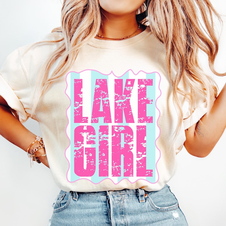 Puede incluir: Camiseta color crema con el texto "LAKE GIRL" en letras rosas desgastadas. El texto est&aacute; sobre un fondo a rayas azul claro y blanco dentro de un borde rosa festoneado. La camiseta se combina con vaqueros azules.