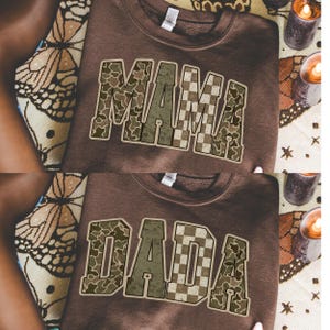 Op de afbeelding: Twee bruine sweatshirts, &eacute;&eacute;n met "MAMA" en de andere met "DADA" in een camouflage- en dambordpatroon. De sweatshirts worden gepresenteerd op een oppervlak met patronen, met kaarsen op de achtergrond.