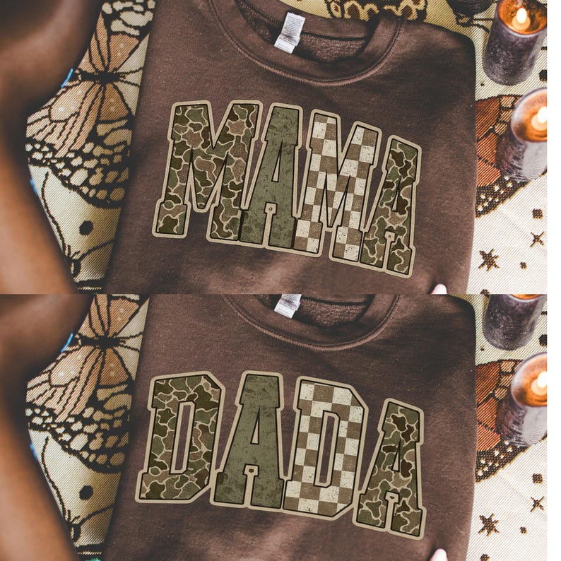 Op de afbeelding: Twee bruine sweatshirts, &eacute;&eacute;n met "MAMA" en de andere met "DADA" in een camouflage- en dambordpatroon. De sweatshirts worden gepresenteerd op een oppervlak met patronen, met kaarsen op de achtergrond.