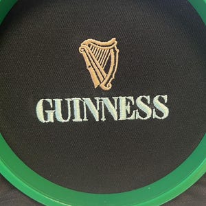 Pode incluir: Um tecido preto com um aro de bordado verde. O aro tem uma harpa bordada em ouro e a palavra "GUINNESS" em branco.