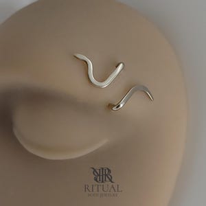 May include: Two silver-toned metal body jewelry pieces with a smooth, curved, snake-like shape. The jewelry is displayed on a neutral-toned surface. Below the jewelry, the text RITUAL BODY JEWELRY is printed in a dark gray, serif font.