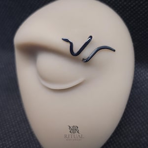Puede incluir: Una joya corporal negra con forma de serpiente colocada sobre un rostro de maniquí beige. La joya presenta un diseño liso y curvado que recuerda a una serpiente. Debajo de la joya, el texto RITUAL BODY JEWELRY está impreso en letras negras.