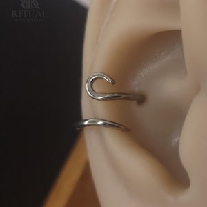 Könnte beinhalten: Nahaufnahme von silbernem Körperschmuck, einem gebogenen Ohr-Cuff und einem kleinen Ring, der an einem Ohr getragen wird. Der Schmuck ist glatt und glänzend. Der Markenname "RITUAL BODY JEWELRY" ist oben links sichtbar.