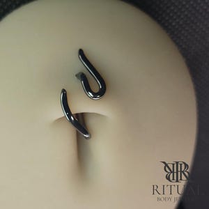 May include: A close-up of a belly button piercing. The piercing is a dark, curved metal design. The jewelry is in the shape of a curved hook. The brand name "RITUAL BODY JEWELRY" is visible in the bottom right corner.