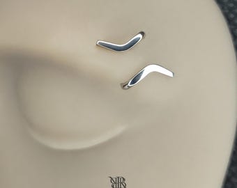 Puede incluir: Dos piezas de joyería corporal plateadas en forma de V están insertadas en una superficie beige lisa. Cada pieza presenta un acabado metálico pulido con una curva angular distintiva. El logo RITUAL BODY JEWELRY aparece impreso abajo.