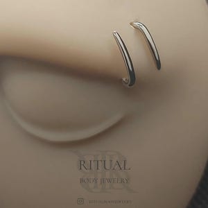 以下が含まれることがあります： ベージュ色の表面に挿入された2つの銀色の湾曲した鼻ピアス。下部には濃いグレーで「RITUAL BODY JEWELRY」という文字が印刷されており、Instagramのアイコンと小さな文字で「RITUALBODYJEWELRY」というハンドルネームが記載されています。