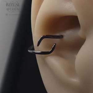 May include: Black body jewelry hoop earring. The earring is a curved, open-ended design. The image shows the earring in an ear piercing. The brand name "RITUAL BODY JEWELRY" is visible in the upper left corner.