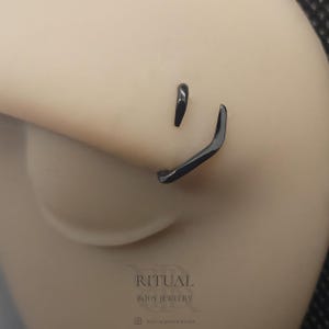 Puede incluir: Primer plano de joyería corporal negra, que incluye un aro de nariz curvado y un pequeño perno. La joyería se coloca sobre un tono de piel neutro. La marca "RITUAL BODY JEWELRY" es visible en la parte inferior de la imagen.