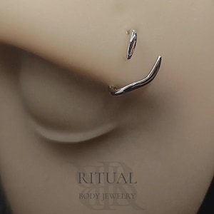 May include: A silver-toned nose ring featuring a curved, wave-like design that follows the contour of the nostril. The jewellery has a polished, metallic finish and is displayed on a neutral skin-toned surface with the text RITUAL BODY JEWELRY.