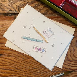 May include: Watercolor illustration of makeup items on a white card. The card features a pencil, eyeliner, eyeshadow, and a small eyeshadow palette. The card is on top of a cream-colored envelope, with a watercolor paint set in the background.