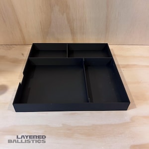 May include: A black, rectangular organizer with multiple compartments. The organizer is divided into sections of varying sizes, suitable for storing small items. The text "LAYERED BALLISTICS" is visible in the lower left corner.