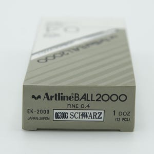 May include: A box of Artline BALL2000 fine point black ink pens. The box is gray with black stripes and the text "Artline BALL2000" in black. The box also has the text "FINE 0.4", "EK-2000", "063003 SCHWARZ", "1 DOZ (12 PCS.)", and "JAPAN/JAPON" in black.