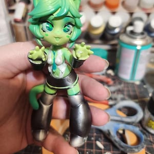 May include: A green and silver painted figurine of a cartoon alien with large eyes and antennas. The figurine is wearing a white and blue outfit with a black skirt and silver boots.