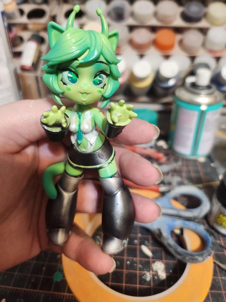 May include: A green and silver painted figurine of a cartoon alien with large eyes and antennas. The figurine is wearing a white and blue outfit with a black skirt and silver boots.