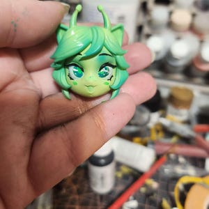 May include: Green and white alien head figurine with large eyes and a toothy grin. The figurine is made of polymer clay and has a textured surface.