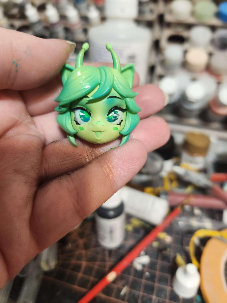 May include: Green and white alien head figurine with large eyes and a toothy grin. The figurine is made of polymer clay and has a textured surface.