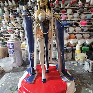 May include: A detailed figurine of a female character with long blonde hair, wearing a blue and white outfit. The figure stands on a red base, with blue accents and gold detailing. The background shows various paint bottles and supplies.