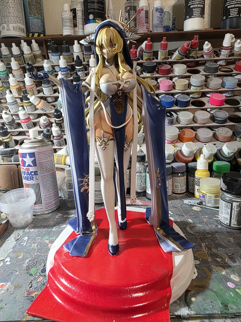 May include: A detailed figurine of a female character with long blonde hair, wearing a blue and white outfit. The figure stands on a red base, with blue accents and gold detailing. The background shows various paint bottles and supplies.