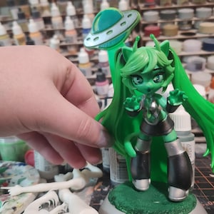May include: Green and silver painted figurine of a female character with long green hair, wearing a black and silver outfit and a silver headband. The figurine is holding her hands up in a gesture of surprise. The figurine is standing on a green base with a silver flying saucer behind it.