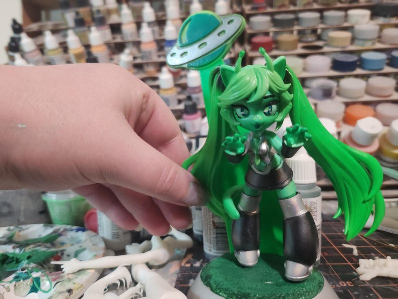 May include: Green and silver painted figurine of a female character with long green hair, wearing a black and silver outfit and a silver headband. The figurine is holding her hands up in a gesture of surprise. The figurine is standing on a green base with a silver flying saucer behind it.