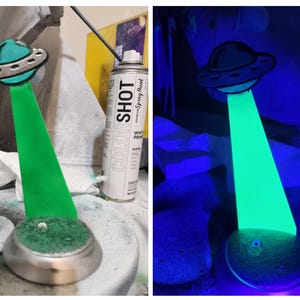 May include: A green glow-in-the-dark UFO figurine with a silver base. The figurine is lit up with a blacklight. The figurine is next to a can of white spray paint.