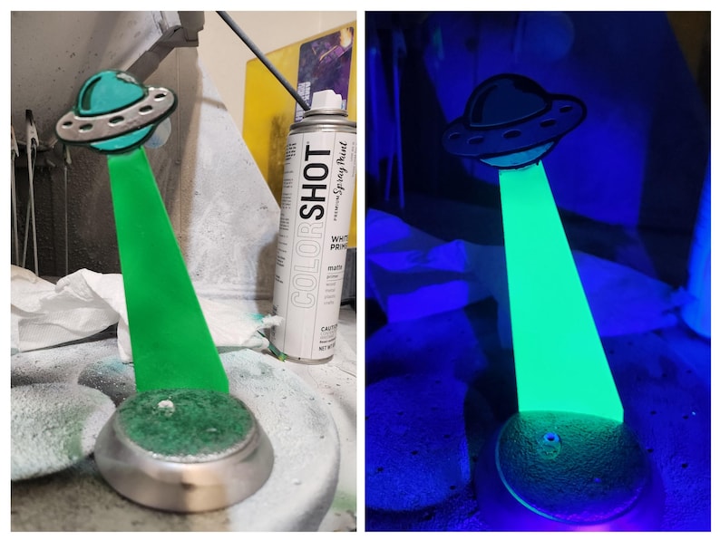 May include: A green glow-in-the-dark UFO figurine with a silver base. The figurine is lit up with a blacklight. The figurine is next to a can of white spray paint.