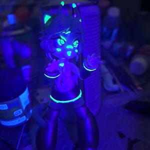 May include: A small, green, blue, and purple figurine of a cartoon character with large eyes and a bright smile. The figurine is glowing under a black light.