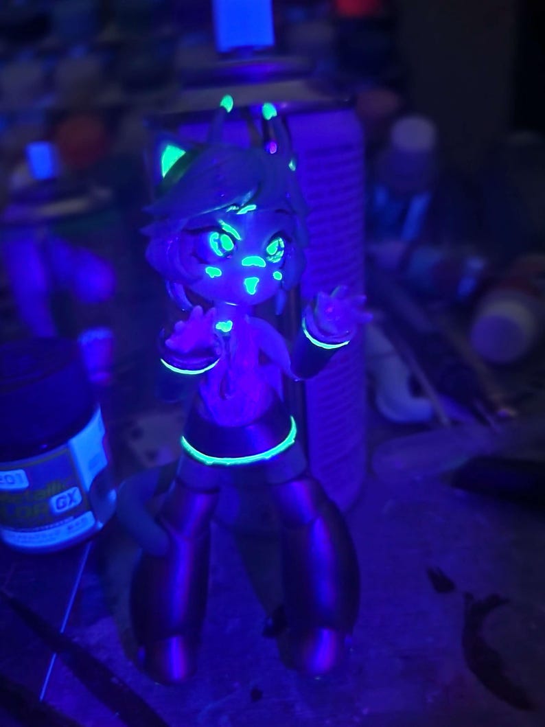 May include: A small, green, blue, and purple figurine of a cartoon character with large eyes and a bright smile. The figurine is glowing under a black light.