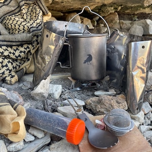 May include: Camping cookware set with a raven logo on a metal pot, a small stove, and a spoon. Includes a tea infuser, a container with an orange cap, and a reflective wind shield. A beige and black patterned scarf is in the background.