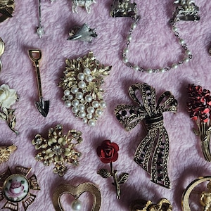 Könnte beinhalten: Eine Sammlung verschiedener Vintage-Broschen und -Anstecknadeln, darunter ein Kreuz, ein Kamel, eine Schaufel, eine Schleife und ein Herz. Die Broschen haben Perlen, Strasssteine und goldfarbenes Metall. Der Hintergrund ist ein rosa Stoff.
