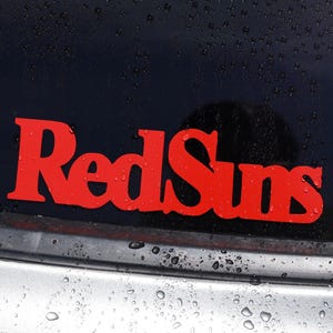 May include: A bright red vinyl decal featuring the text RedSuns in a bold, stylized font. The decal is applied to the dark, wet glass surface of a vehicle window, with visible water droplets scattered across the dark background.