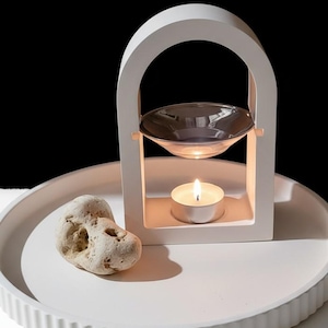 Könnte beinhalten: Ein weißer Aromatherapie-Öldiffusor mit einer durchsichtigen Glasschale, einer brennenden Teelichtkerze und einem dekorativen Stein auf einem weißen Tablett. Der Diffusor hat ein bogenförmiges Design und ist vor einem schwarz-weißen Hintergrund platziert.