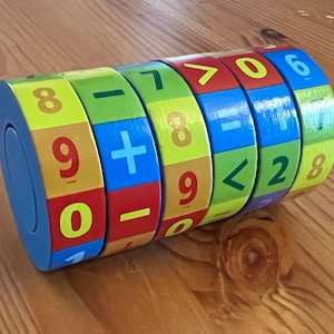 Può includere: Un giocattolo di legno colorato con cilindri rotanti che presentano numeri, simboli matematici e segni maggiore di e minore di. Il giocattolo è progettato per aiutare i bambini ad imparare i concetti matematici di base.