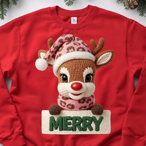 Puede incluir: Sudadera roja con un diseño de reno de ganchillo, con un gorro y bufanda con estampado de leopardo rosa y marrón. El reno sostiene un letrero con la palabra "MERRY". Ropa navideña.