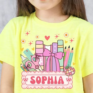 Puede incluir: Una camiseta amarilla brillante con un diseño temático escolar. El gráfico incluye libros, lápices y el nombre "SOPHIA" en un marco decorativo. El diseño es colorido y adecuado para niños.