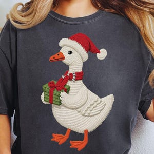 May include: Dark gray t-shirt featuring a whimsical goose illustration. The goose wears a Santa hat, a red scarf, and holds a green gift box with a red ribbon. The design is festive and perfect for the holiday season.