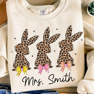 Puede incluir: Sudadera color crema con tres siluetas de conejos con estampado de leopardo, cada una con un lazo amarillo, rosa y melocotón. El texto "Mrs. Smith" está impreso en escritura negra debajo de los conejos. También se incluyen pequeñas estrellas negras.