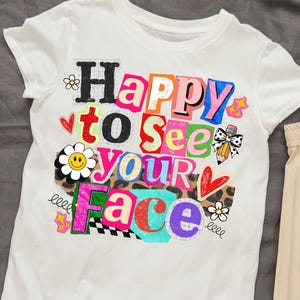 Puede incluir: Camiseta blanca con la frase "Happy to See Your Face" en un diseño colorido estilo collage. El diseño incluye corazones, flores, estrellas y un lápiz con un lazo. La camiseta está hecha de un material suave.