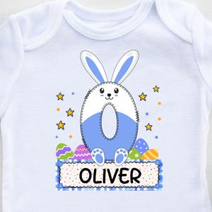 Puede incluir: Body de bebé blanco con un diseño de conejito azul y blanco. El conejito tiene forma de número cero, con el nombre "OLIVER" impreso debajo. Huevos de Pascua y estrellas decoran el diseño.