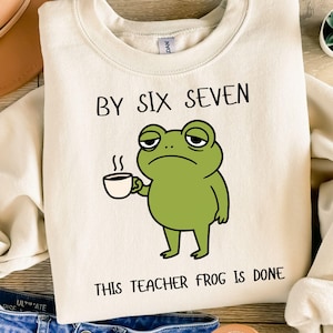 Puede incluir: Sudadera color crema con un gráfico de una rana verde cansada sosteniendo una taza de café. El texto "BY SIX SEVEN" y "THIS TEACHER FROG IS DONE" está impreso en negro. La sudadera está doblada sobre una superficie de madera.