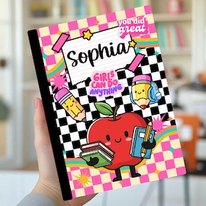 Puede incluir: Un cuaderno con fondo de cuadros blancos y negros y bordes de cuadros rosas y amarillos. La portada muestra el nombre Sophia, el texto you did great y girls can do anything, junto a una manzana de dibujos animados sosteniendo libros y lápices.